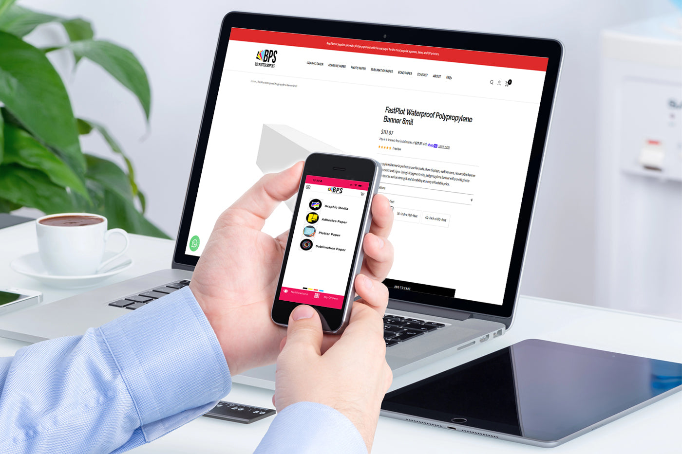 Person using a smartphone with a website displayed on a laptop in an office setting.